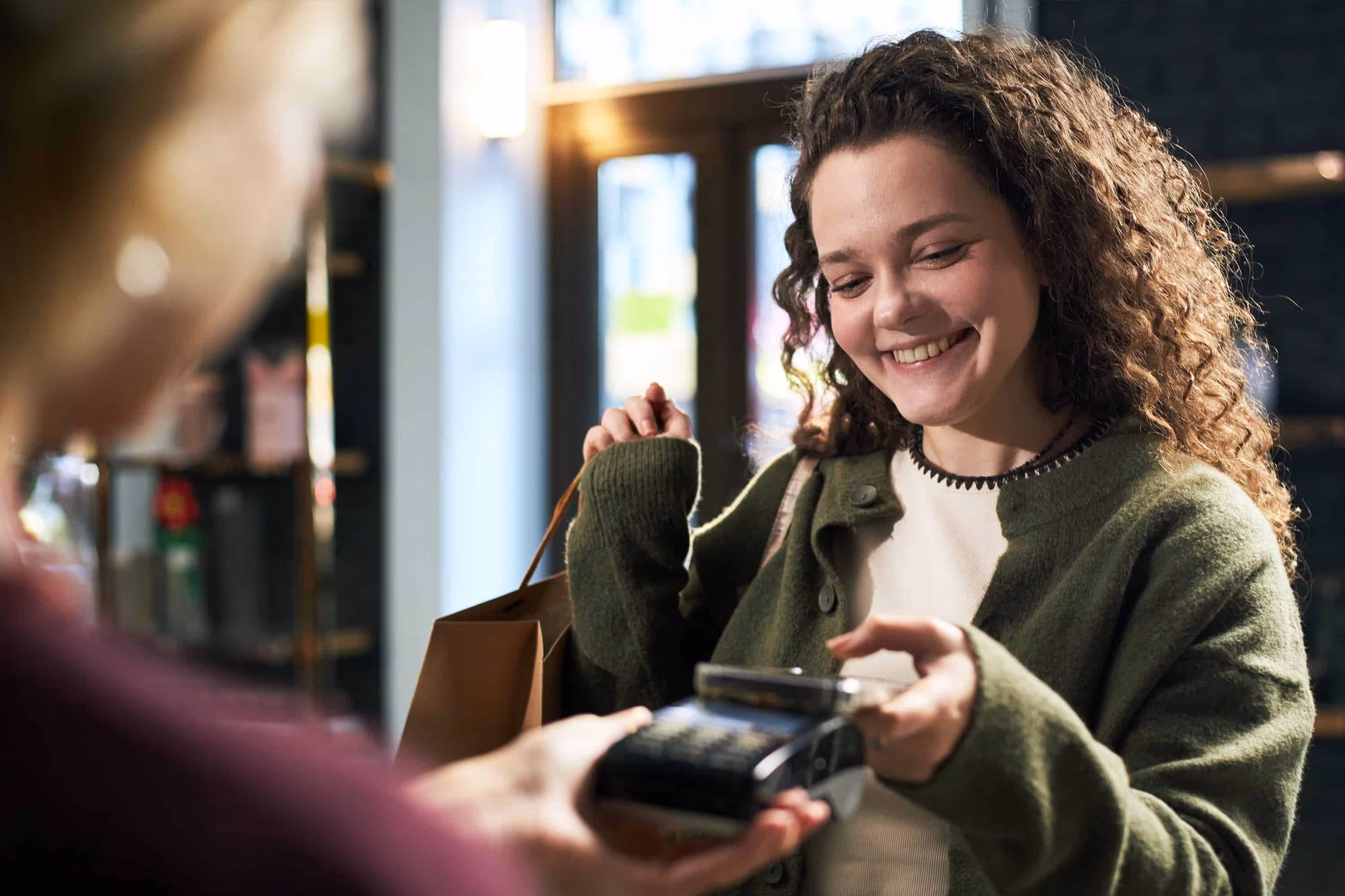 Smiling woman in green sweater paying with phone using a contactless card reader while holding shopping bag.