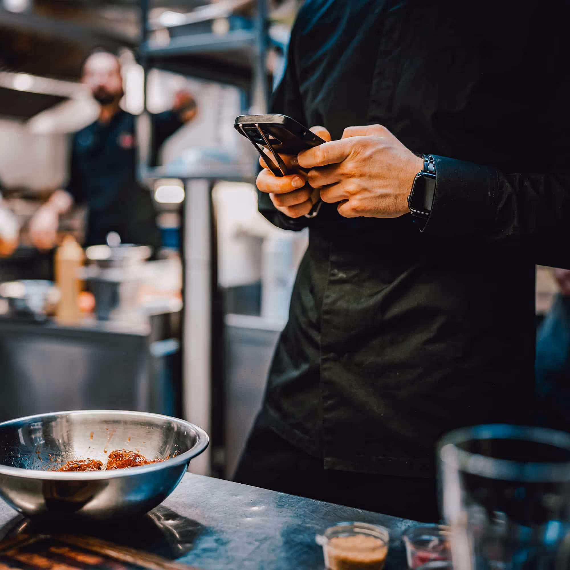 Chef in black uniform using smartphone in a kitchen near a metal bowl with food.