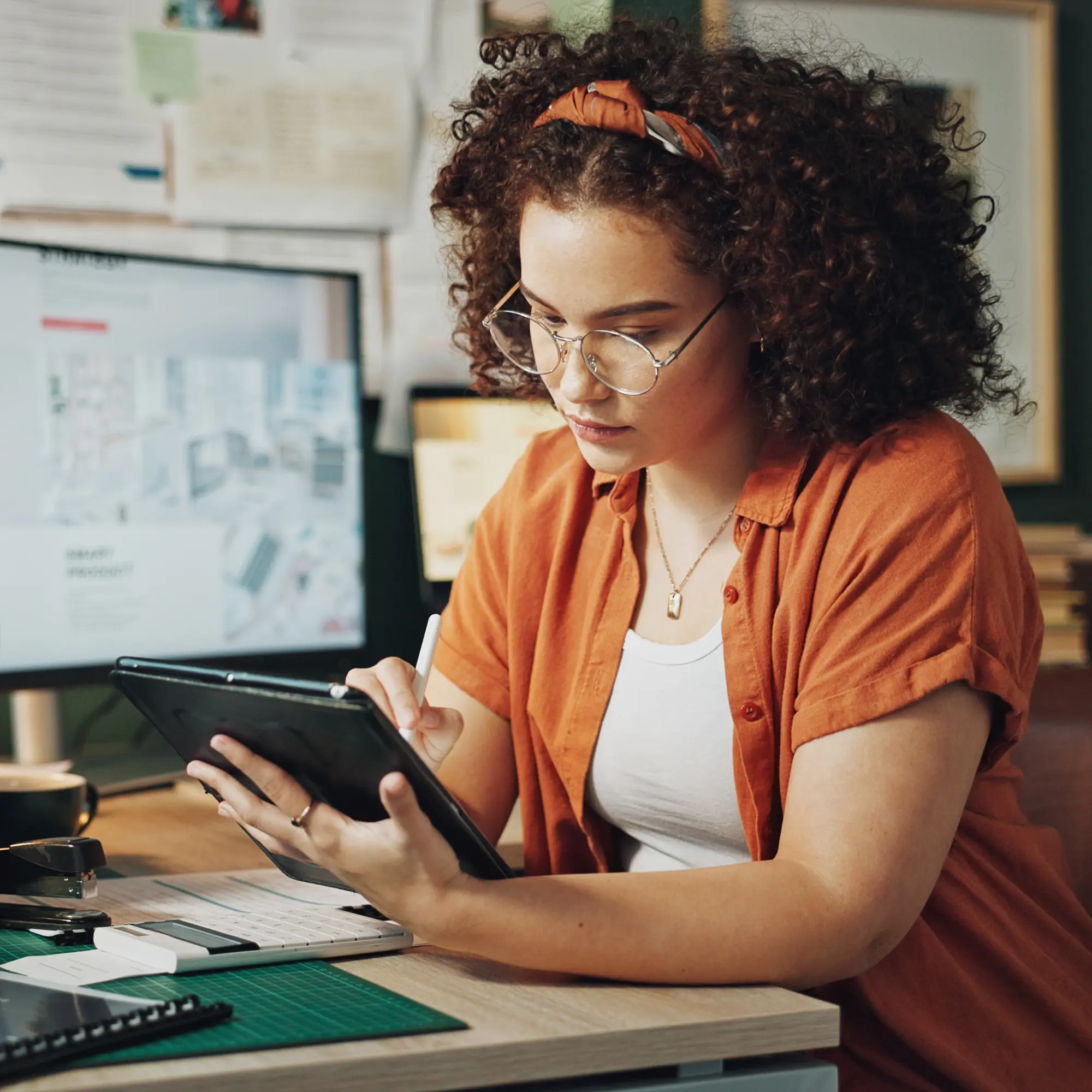 Woman with curly hair and glasses using a stylus on a tablet at a desk with a computer monitor.