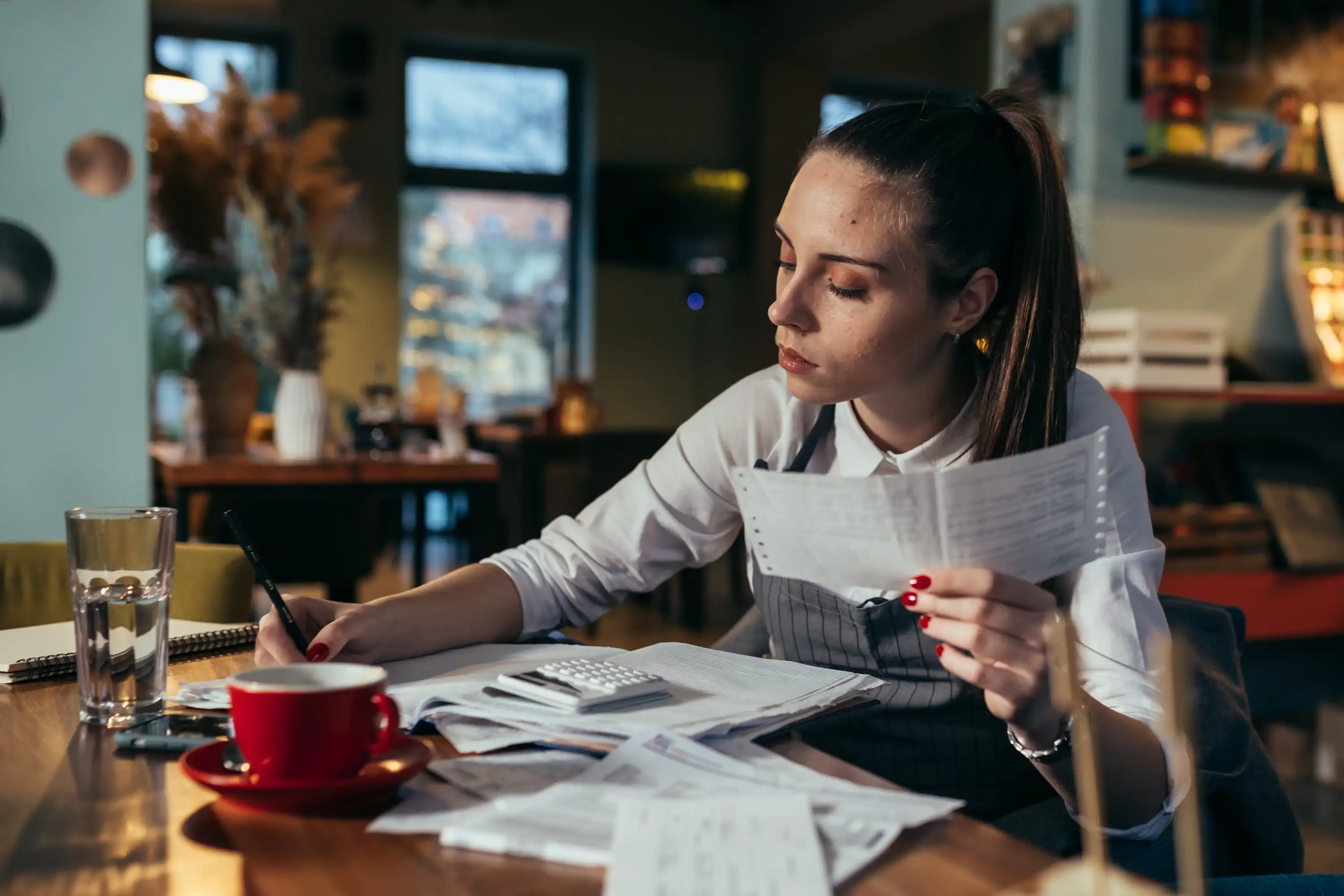 Woman working on finances at a table with papers, calculator, red cup, and glass of water.