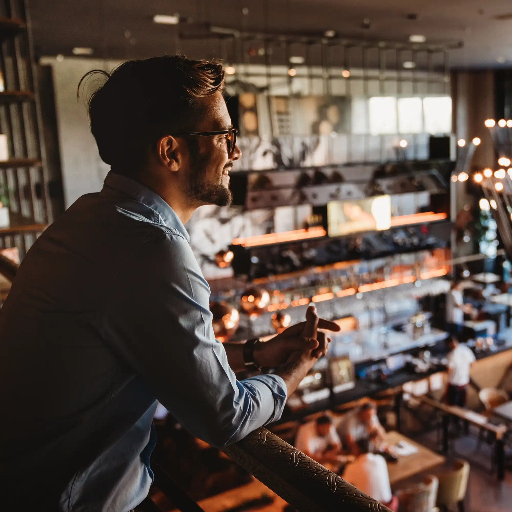 Man with glasses leaning on railing smiling inside a warmly lit cafe or restaurant.