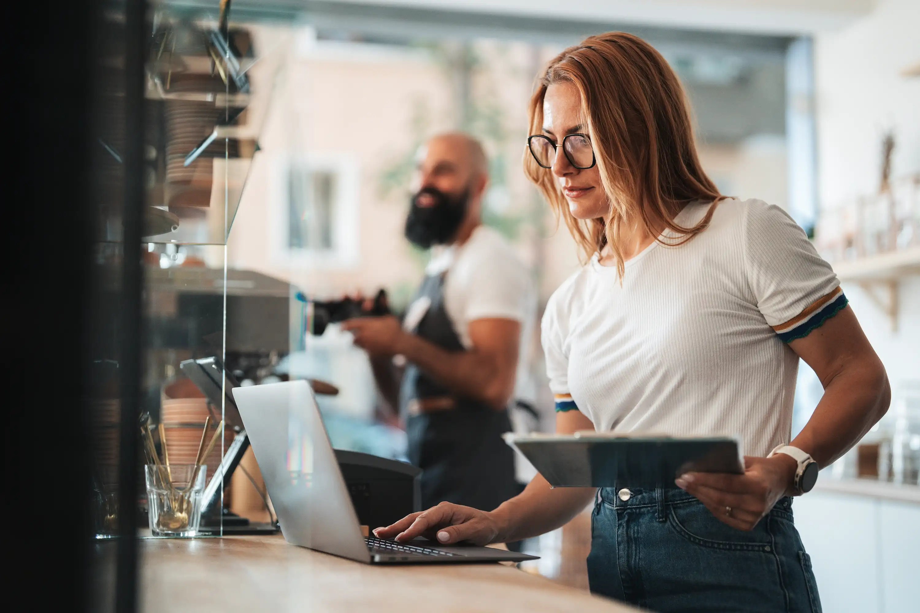 Woman using laptop and holding tablet at cafe counter with barista working in background.