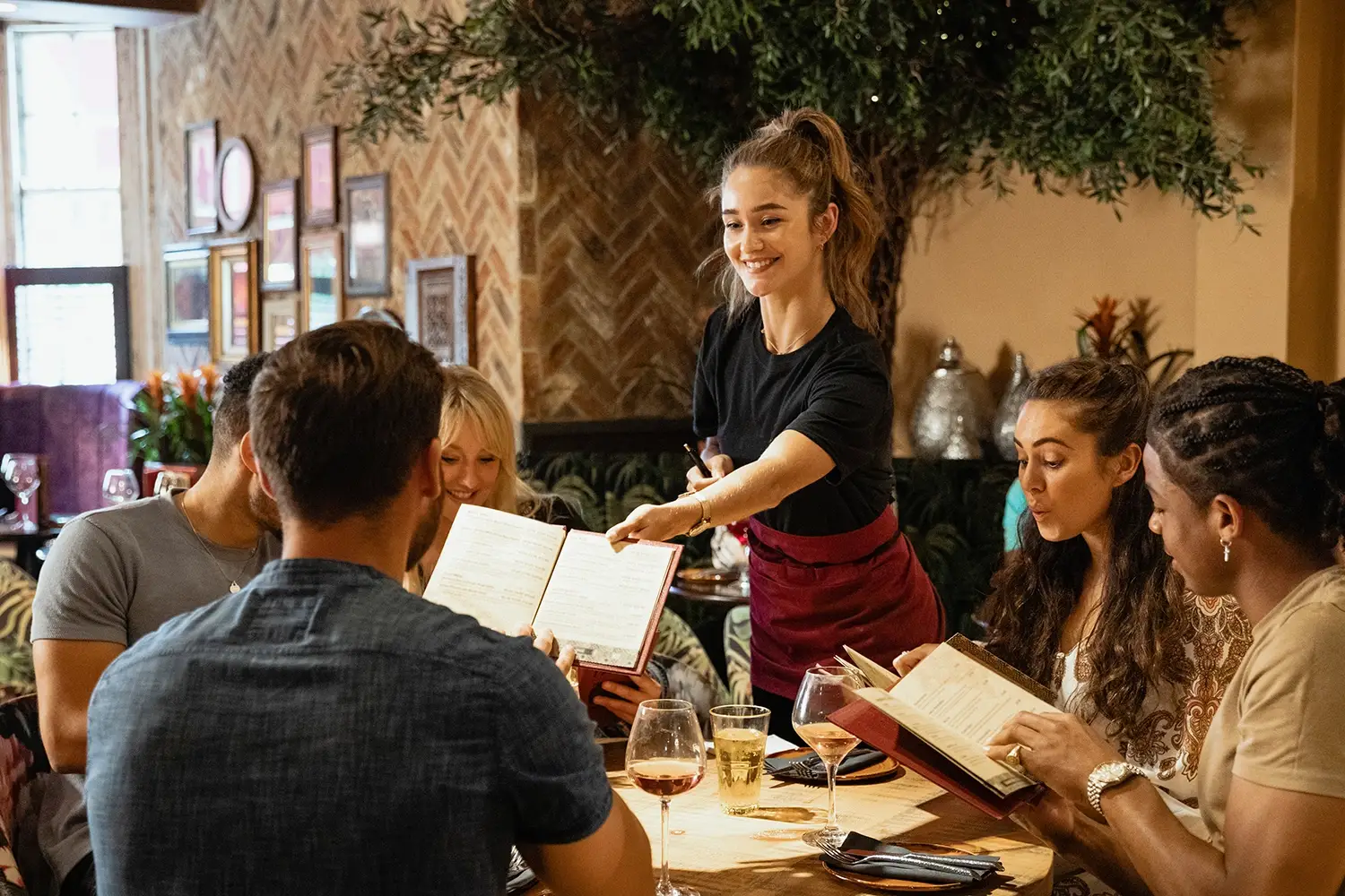 Smiling waitress takes order from a group of friends looking at menus in a cozy restaurant.