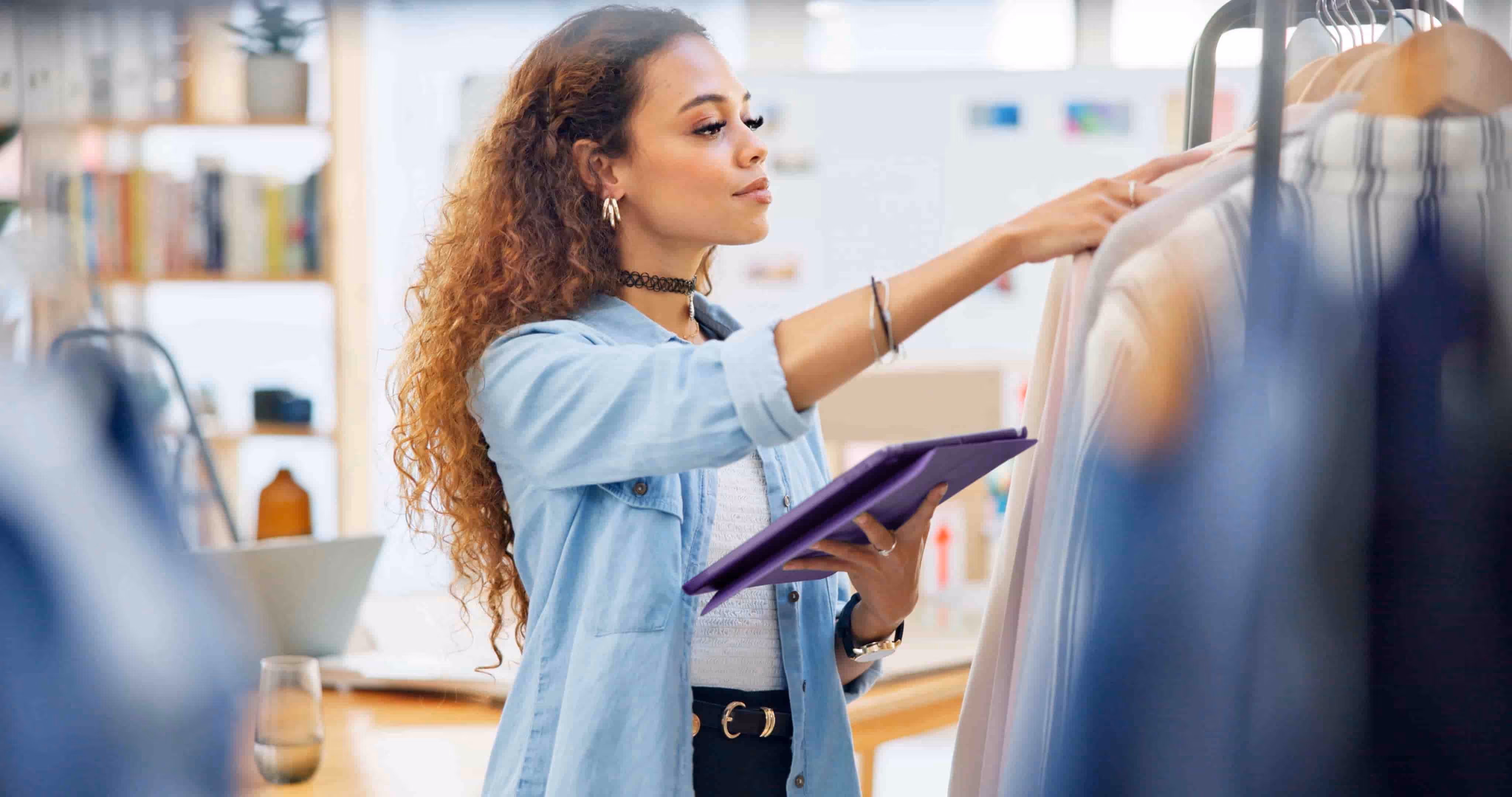Woman in denim shirt holding a tablet and selecting clothes on a rack in a bright room.