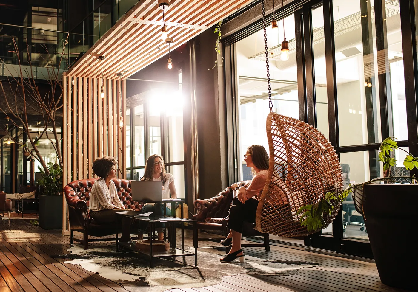 Three women chatting in a modern office lounge, one seated on a hanging wicker chair.