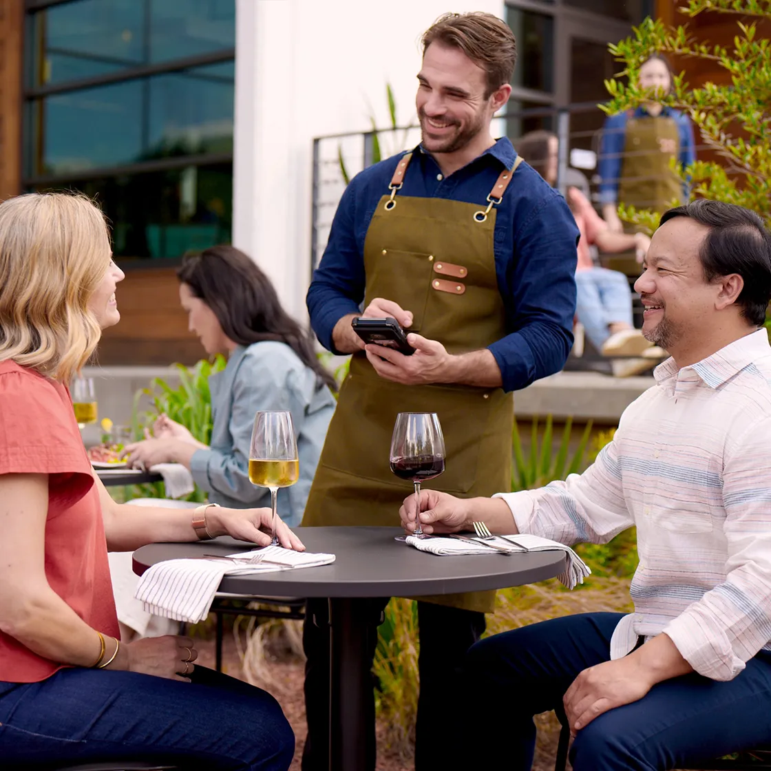 A waiter smiling while taking orders from a man and a woman sitting at an outdoor table with wine glasses.