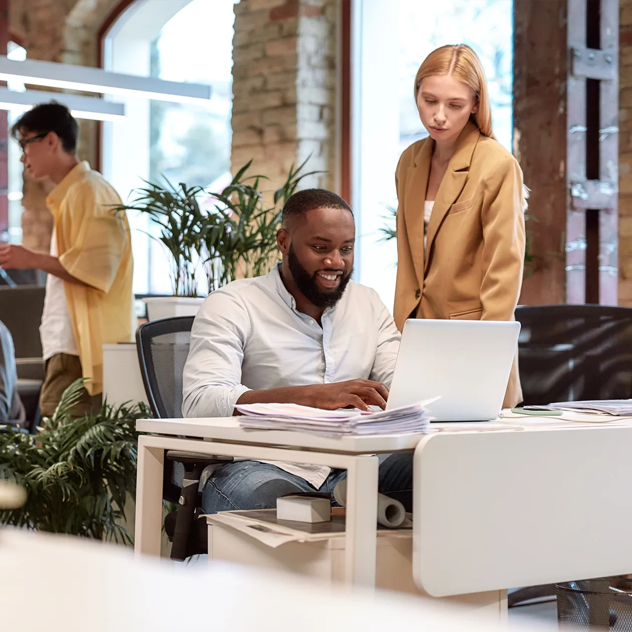 Smiling man sitting at desk working on laptop with woman standing beside him in modern office.
