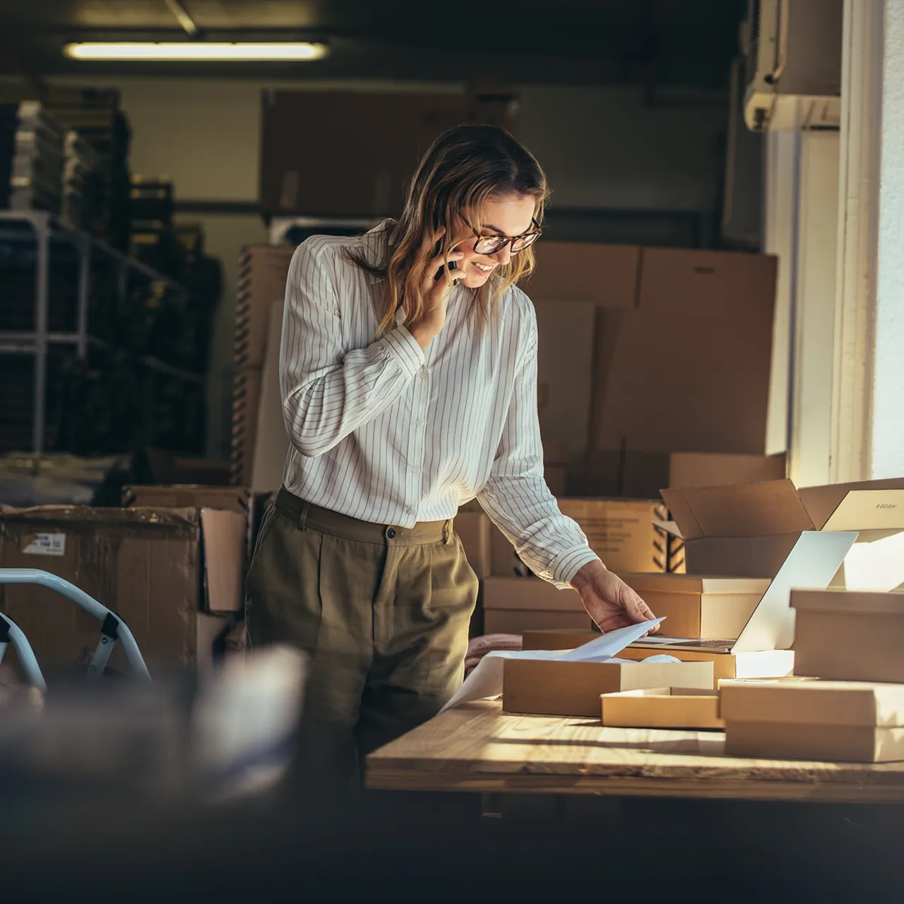 Woman in a warehouse packing boxes while talking on the phone and looking at documents.