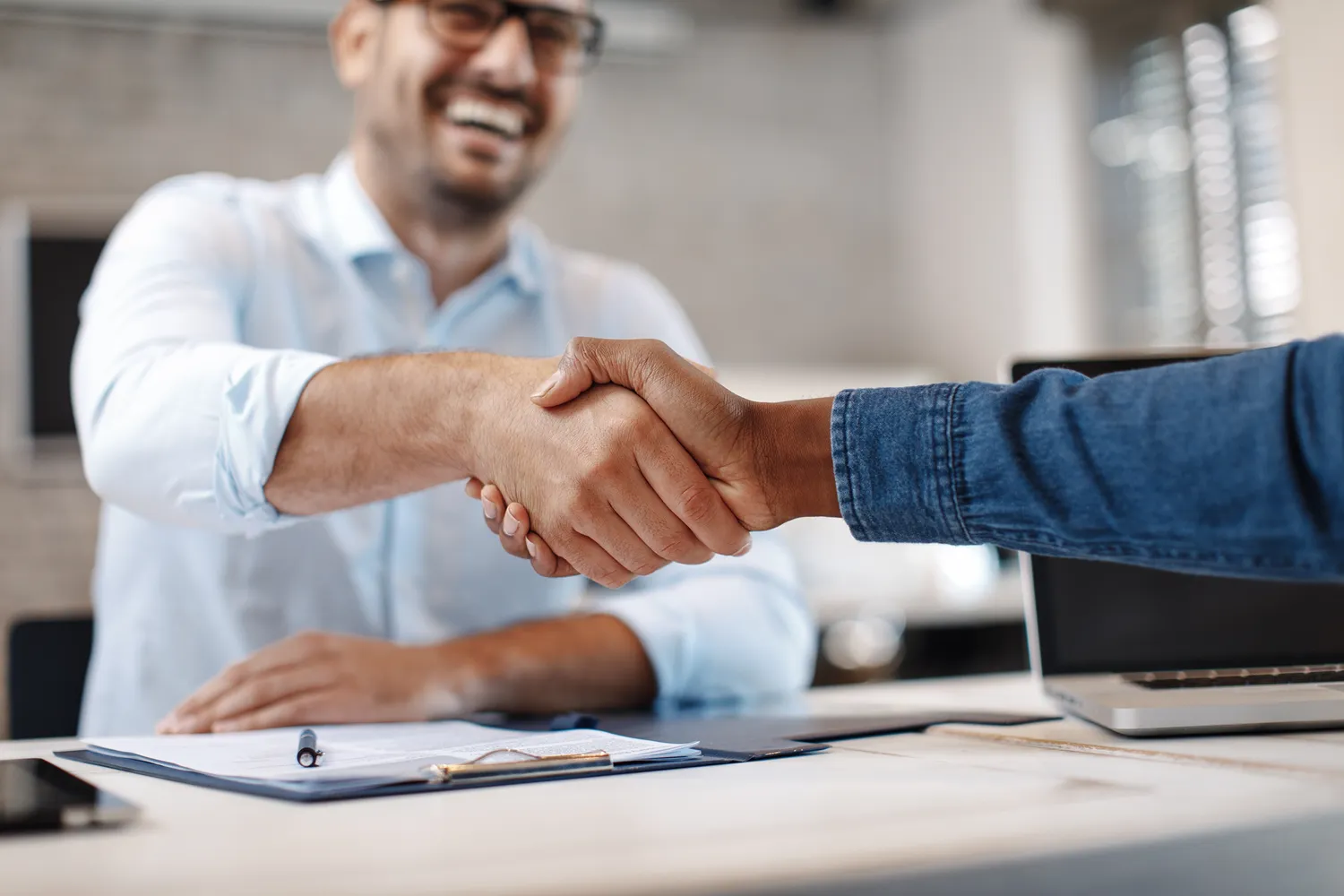 Two people shaking hands across a desk with documents and a laptop.