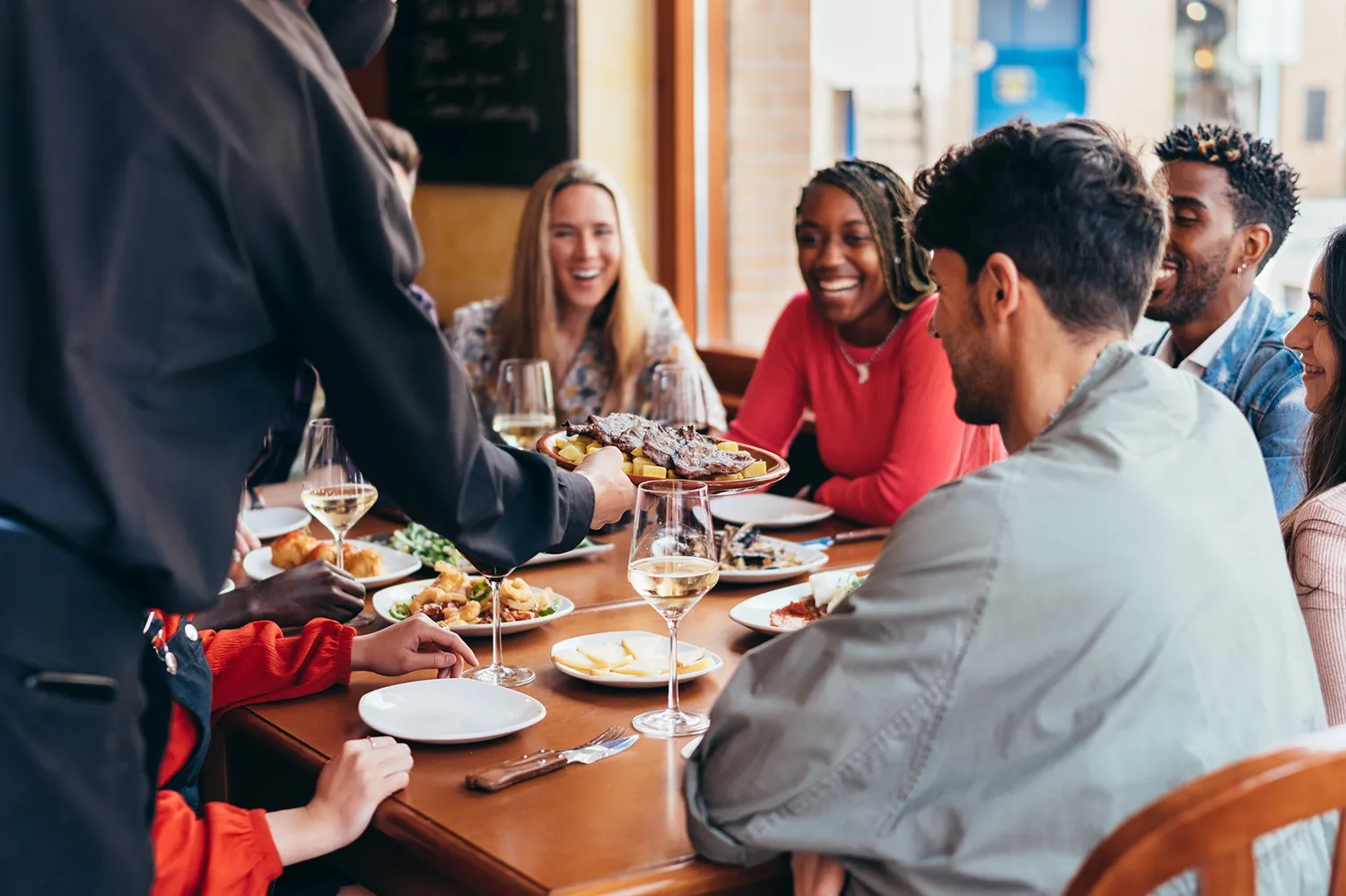 Server serving steak and fries to a group of friends dining together with wine glasses on the table.