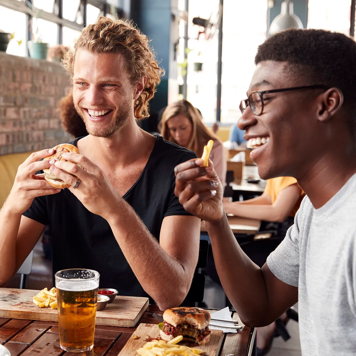 Two men happily eating burgers and fries together at a restaurant table with a glass of beer.