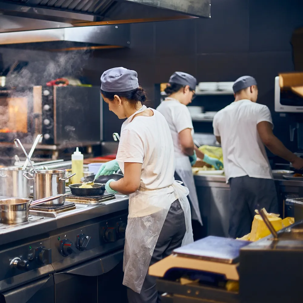 Three chefs wearing white shirts and gray hats cooking together in a busy commercial kitchen.