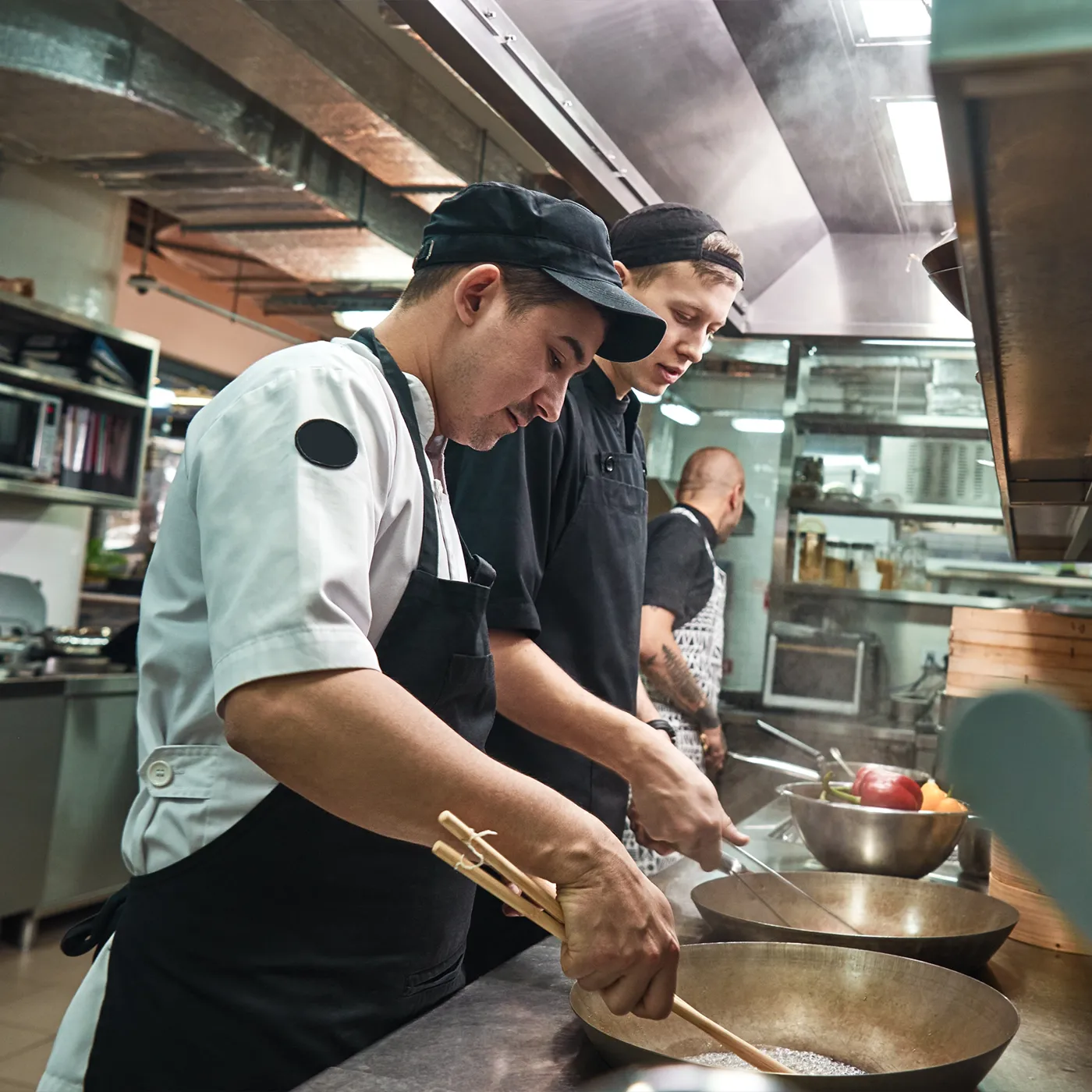 Two chefs cooking in a professional kitchen using large woks while a third chef works in the background.