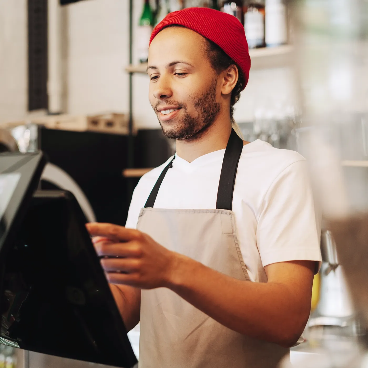 Barista wearing red beanie and apron using touchscreen at coffee shop counter.