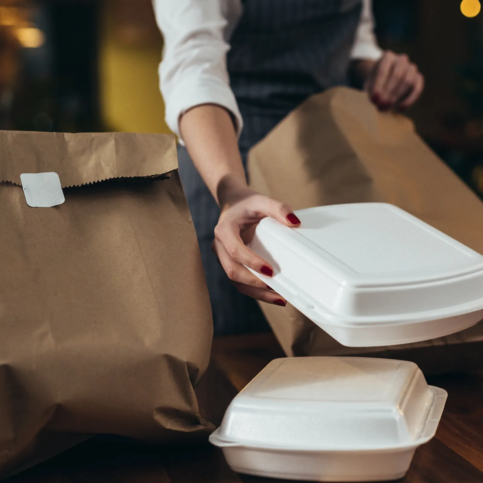 Person packing white styrofoam food containers and brown paper bags on a wooden surface.