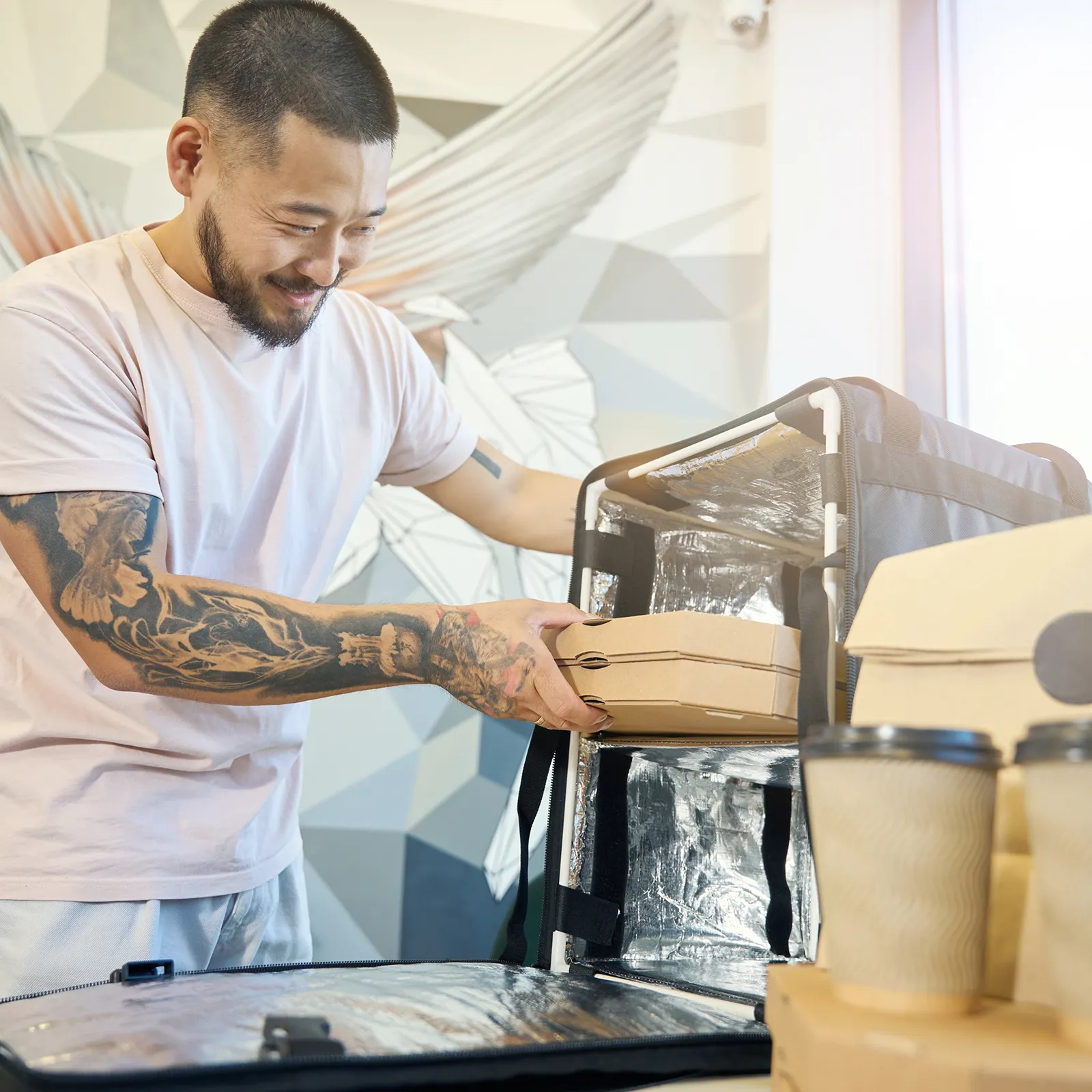 Smiling man with tattoos placing pizza boxes in an insulated food delivery bag.