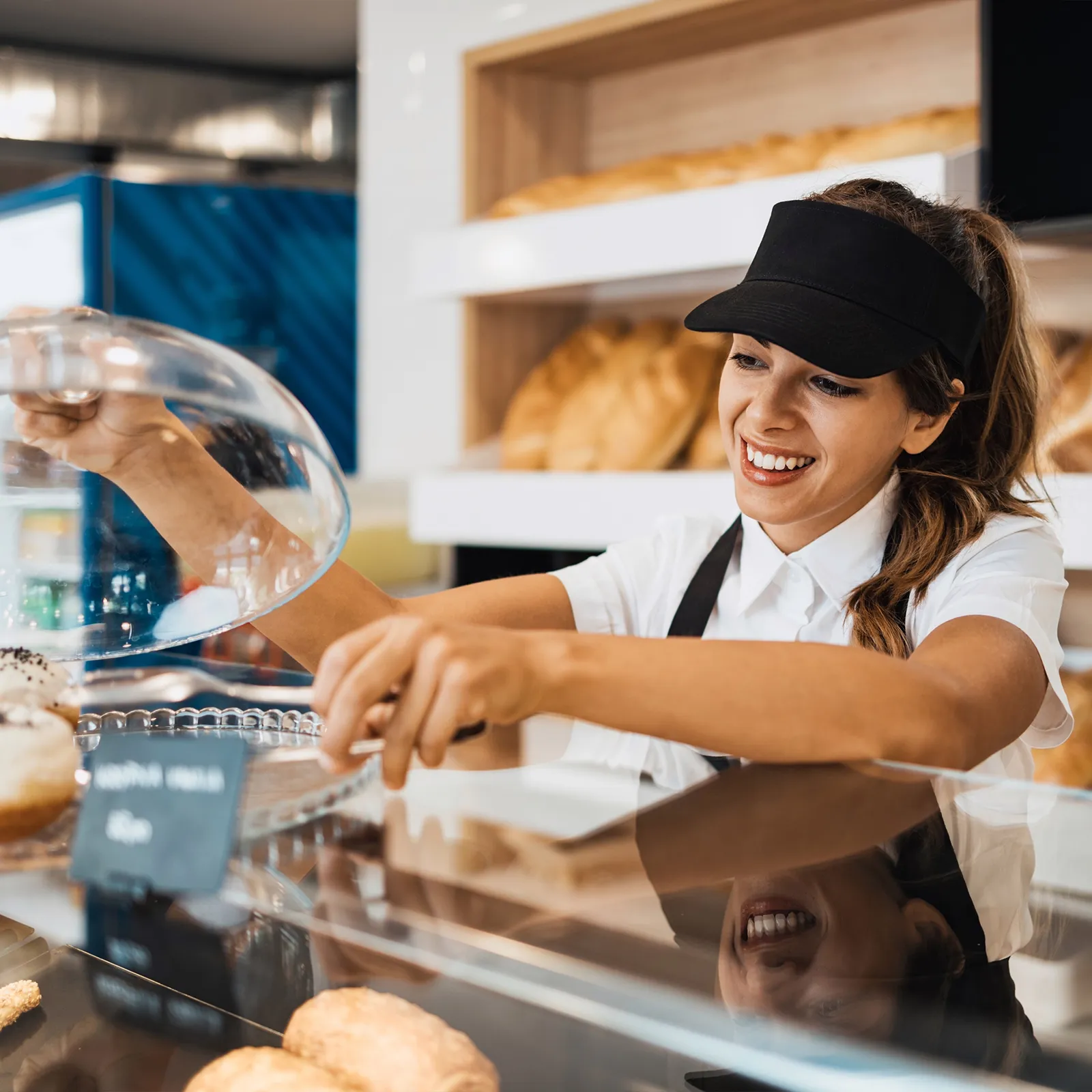 Female bakery worker smiling while lifting a glass cover from pastries behind the counter.