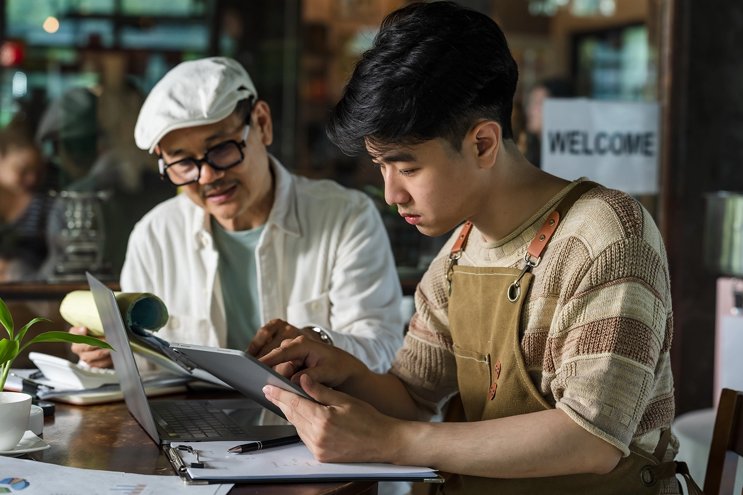 Young man in apron using a tablet while an older man watches, with laptop and papers on table.