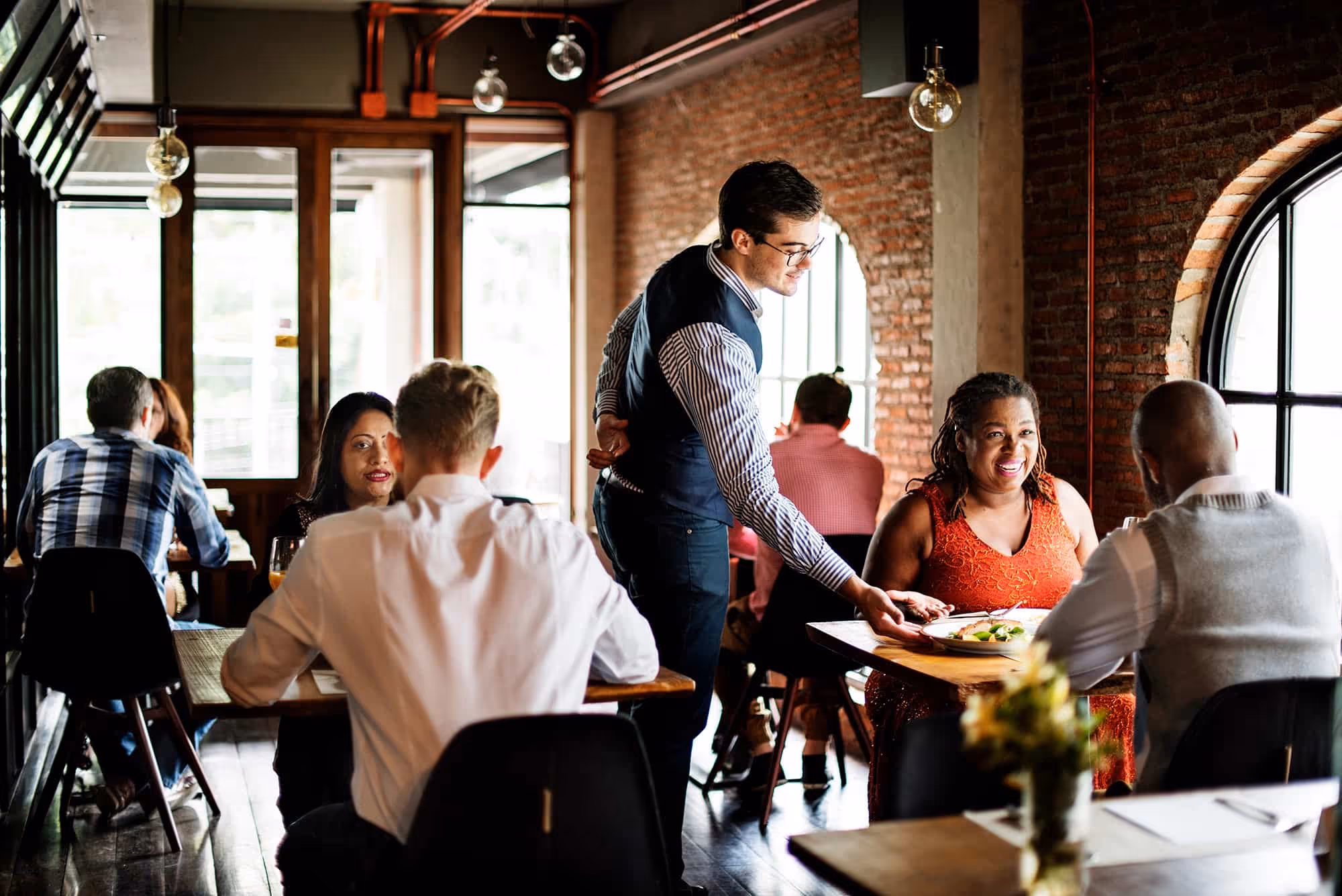 A waiter serves food to a woman smiling in a busy restaurant with several seated diners.