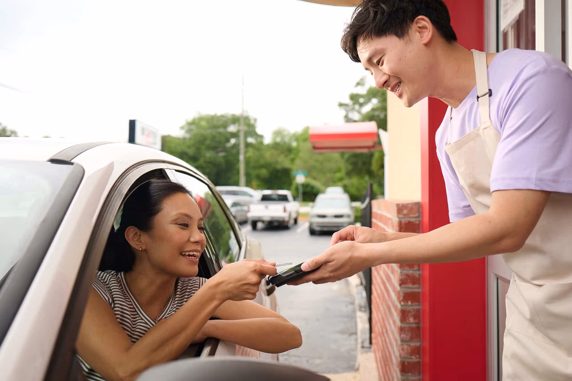 Smiling woman in car paying with card to man in apron at drive-thru window.