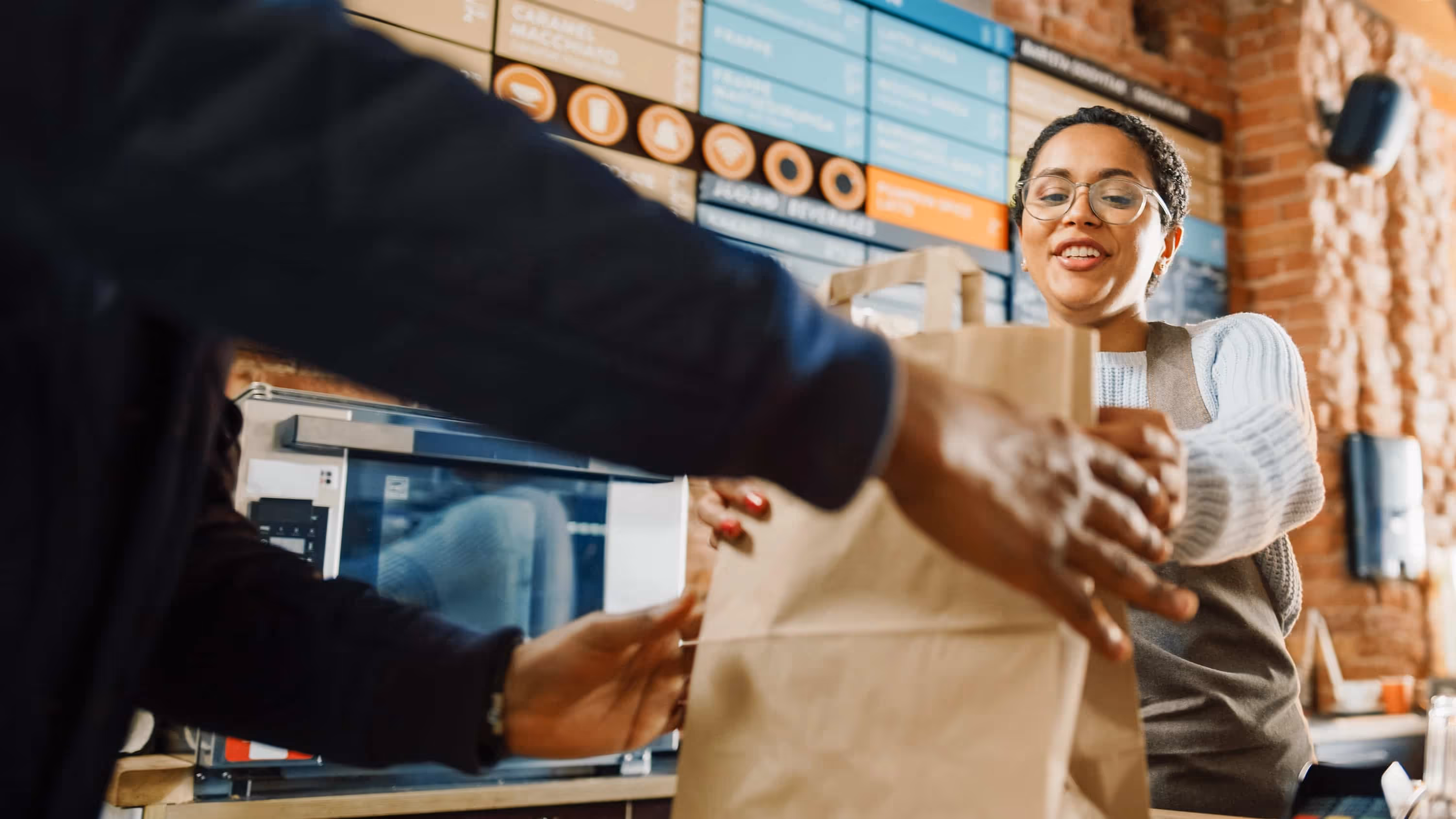 Smiling café worker handing a paper bag order to a customer across the counter.