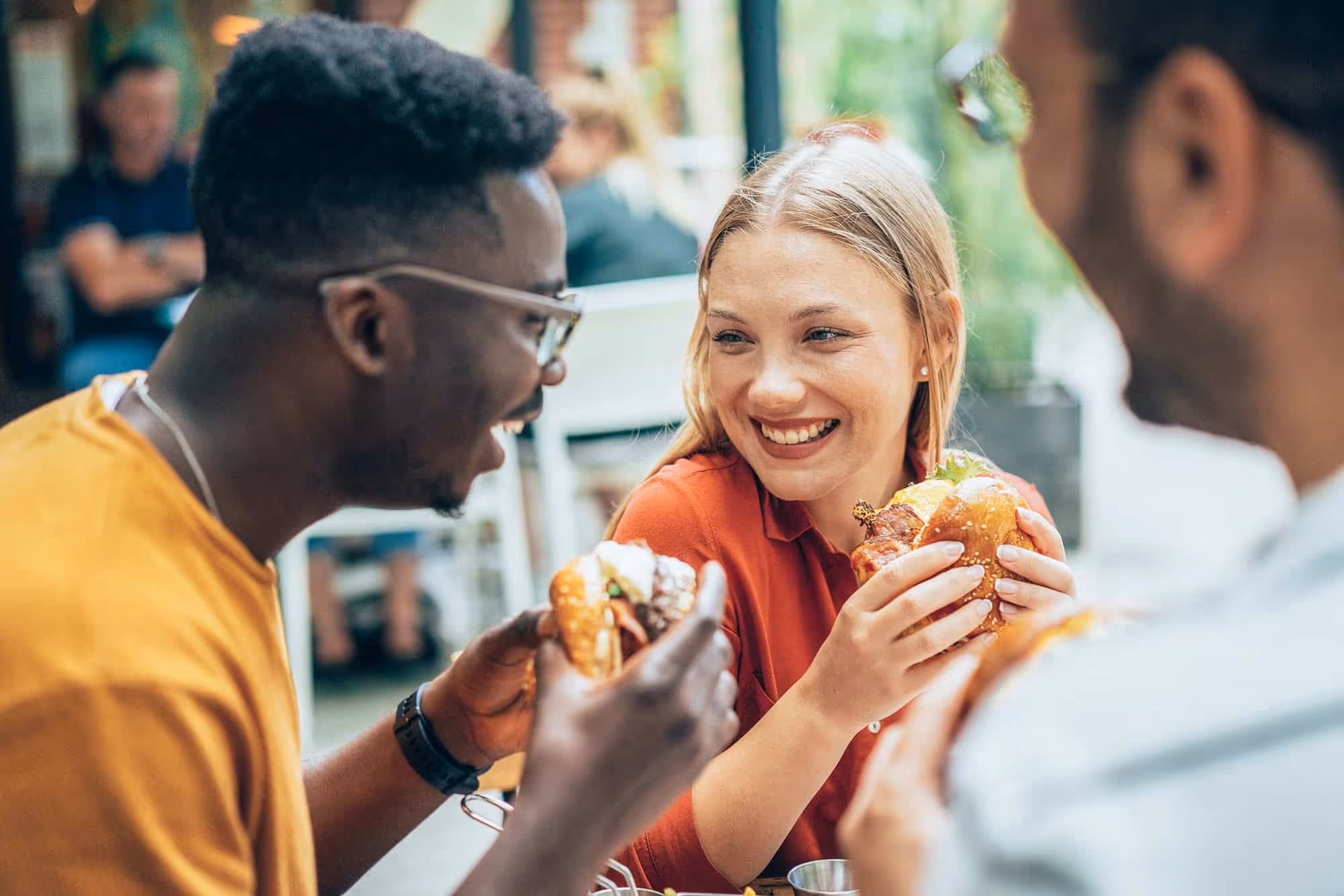 Three friends smiling and holding burgers while enjoying a meal together outdoors.