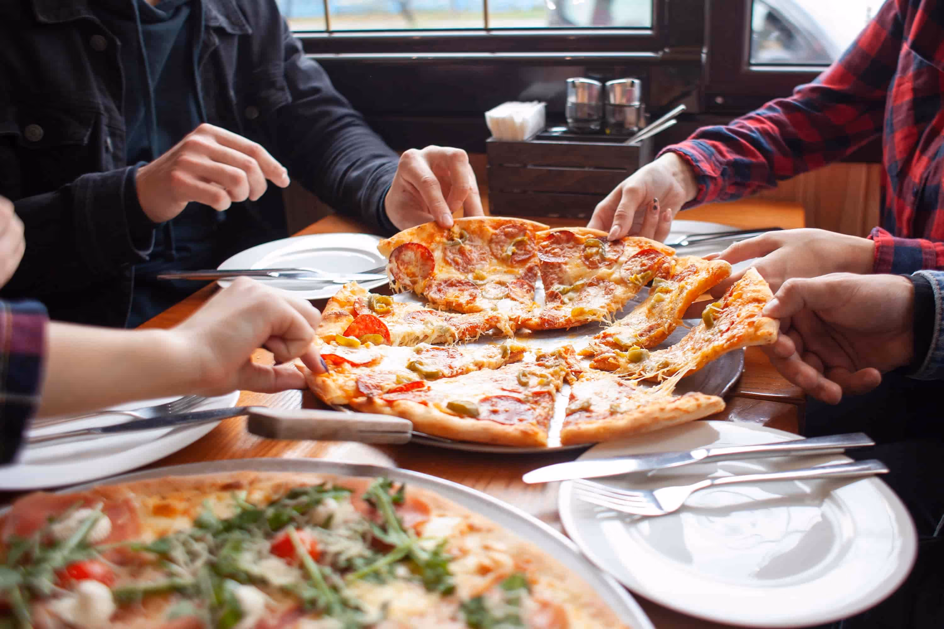 Four hands grabbing slices of pepperoni and olive pizza from a wooden table at a restaurant.