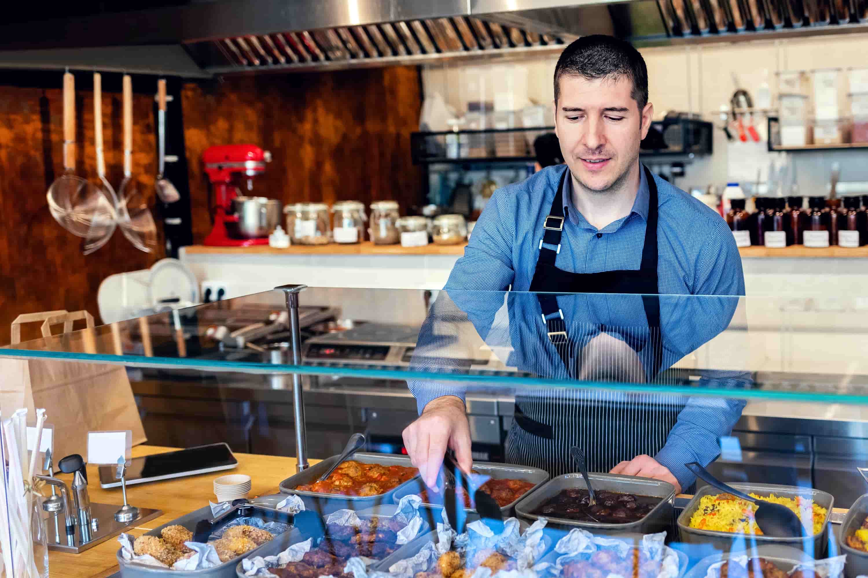 Man in apron serving food with tongs behind a glass counter with various dishes in a restaurant kitchen.