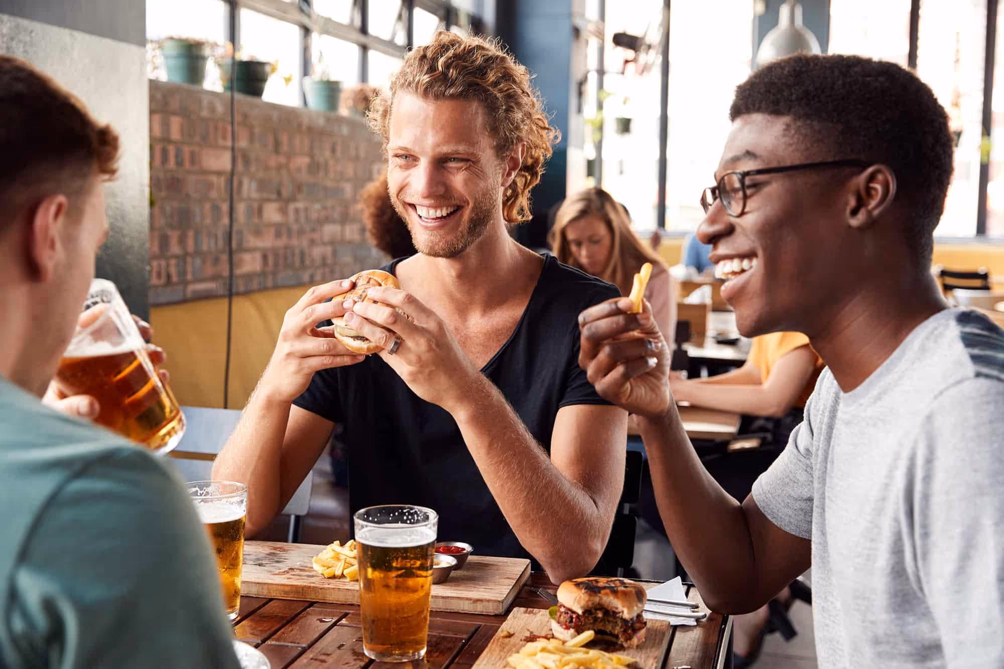 Three men laughing and eating burgers and fries with glasses of beer at a restaurant table.