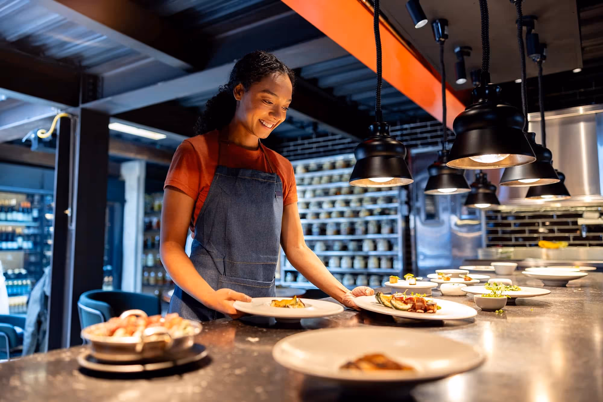 Smiling waitress wearing apron arranging plates of food on restaurant counter under hanging lights.