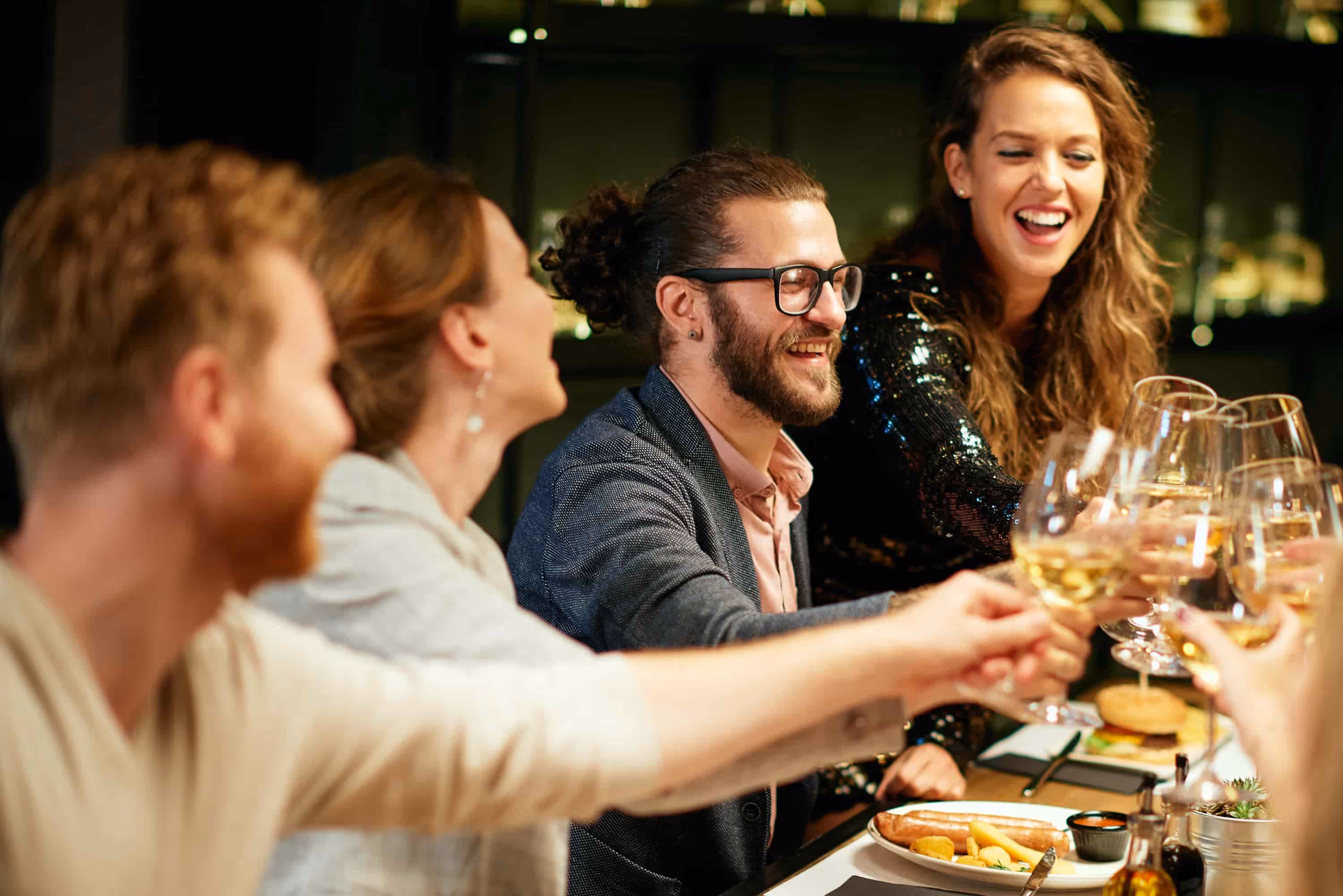 Group of friends smiling and toasting with white wine glasses at a dinner table with food.