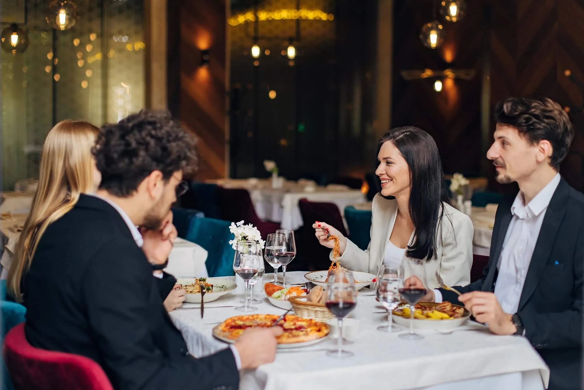 Four people dressed formally enjoying a meal with pizza, pasta, and wine at a restaurant table.