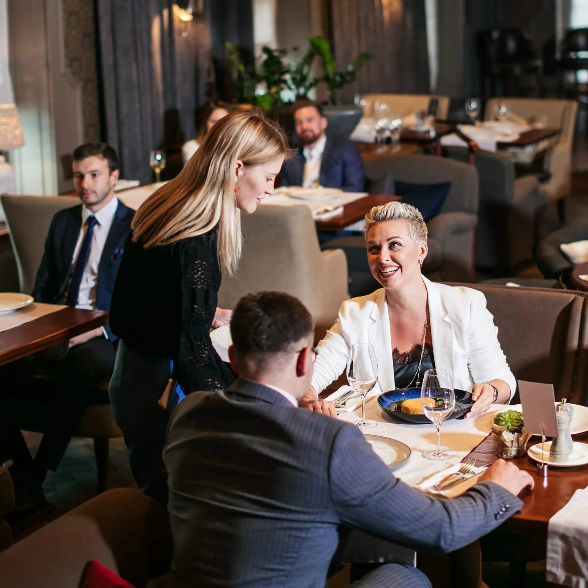 Woman in white jacket smiling at a restaurant table while a server takes her order.