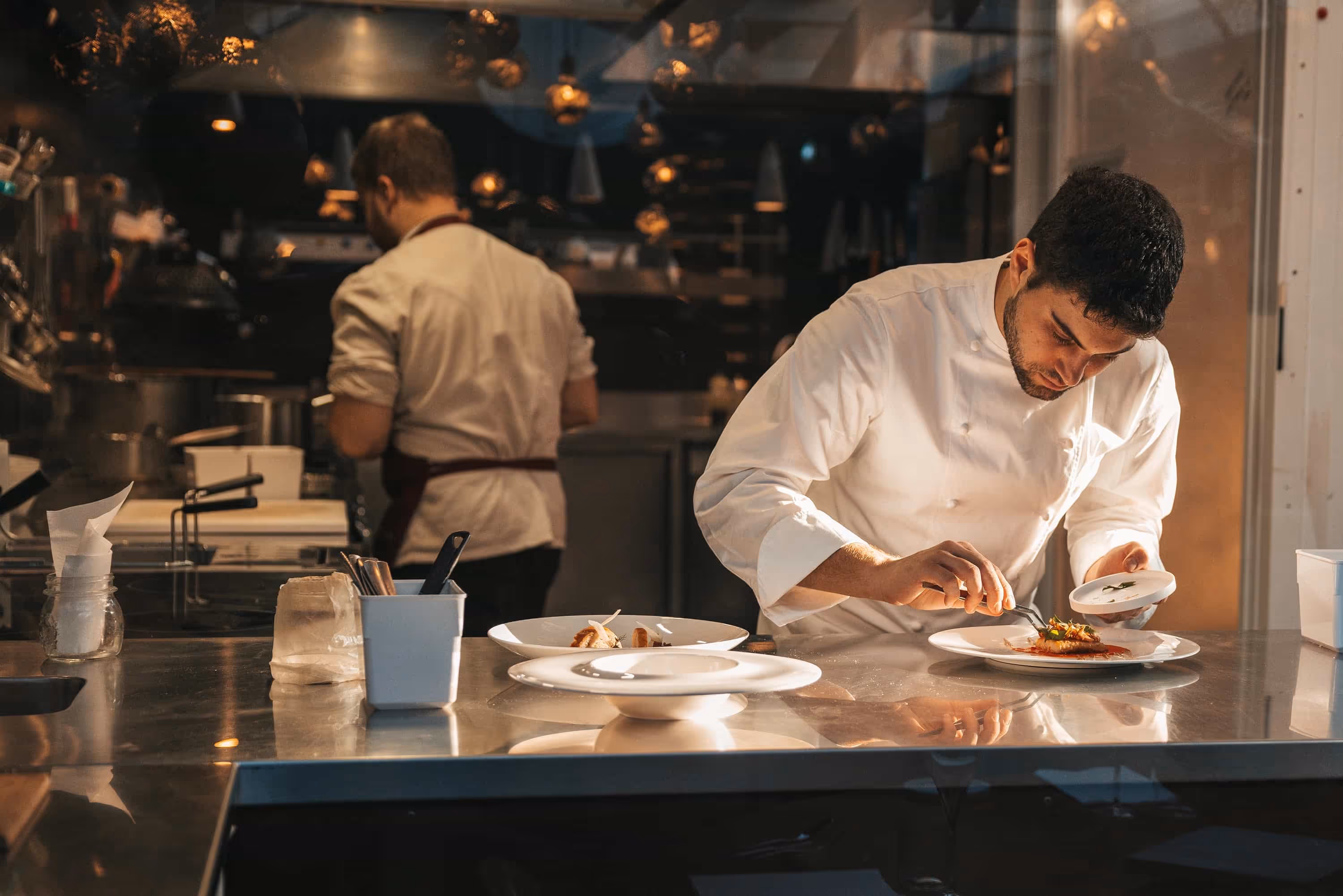 Chef in white coat carefully plating a gourmet dish in a professional kitchen at sunset.