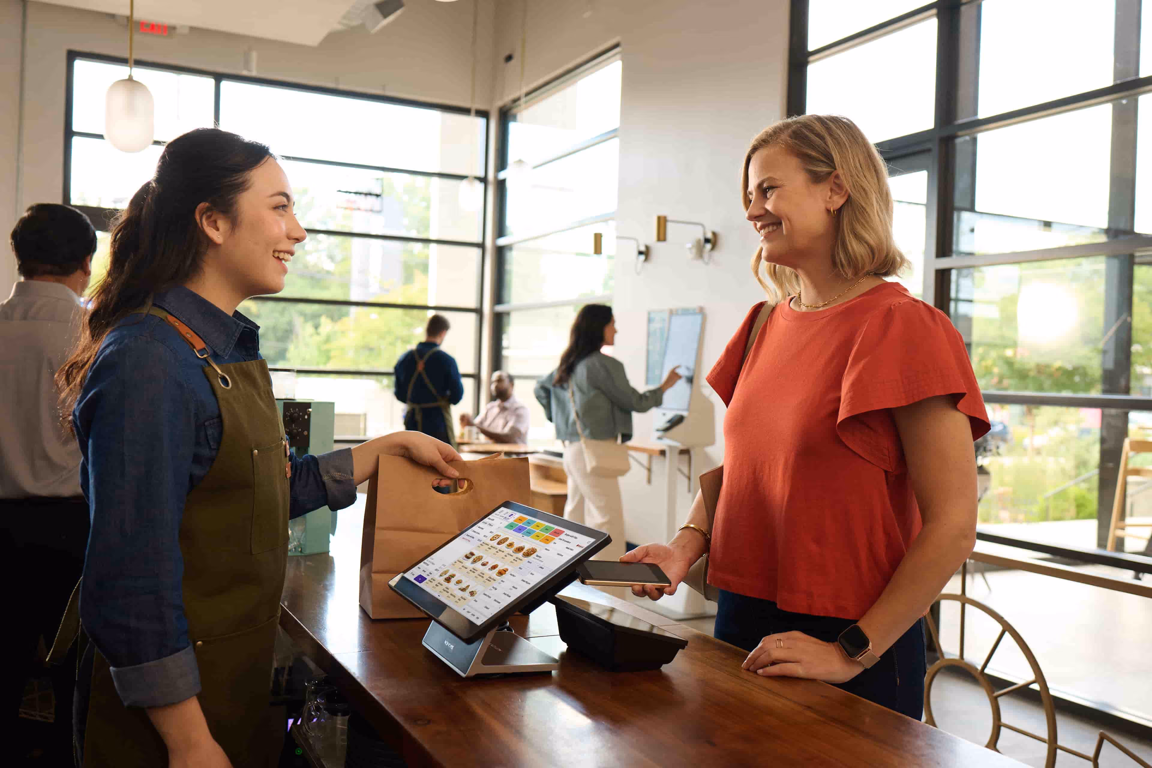 Barista handing a paper bag to a smiling customer paying with smartphone in a bright cafe.
