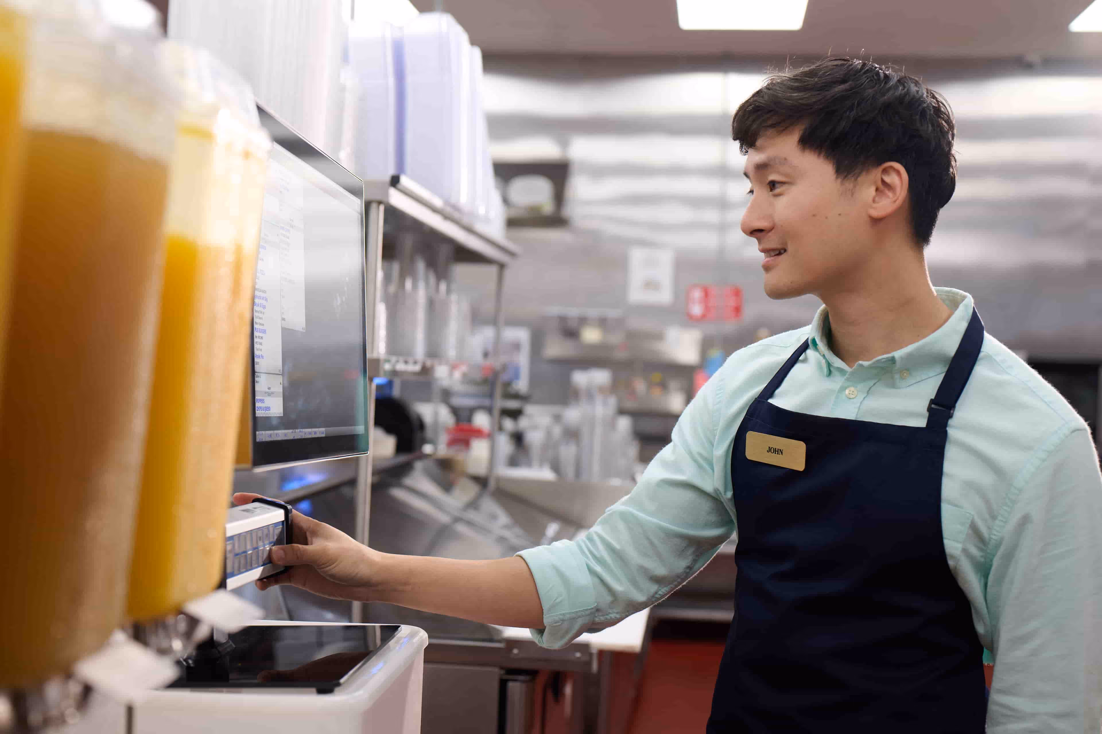 Man in apron labeled John using a touchscreen in a commercial kitchen.