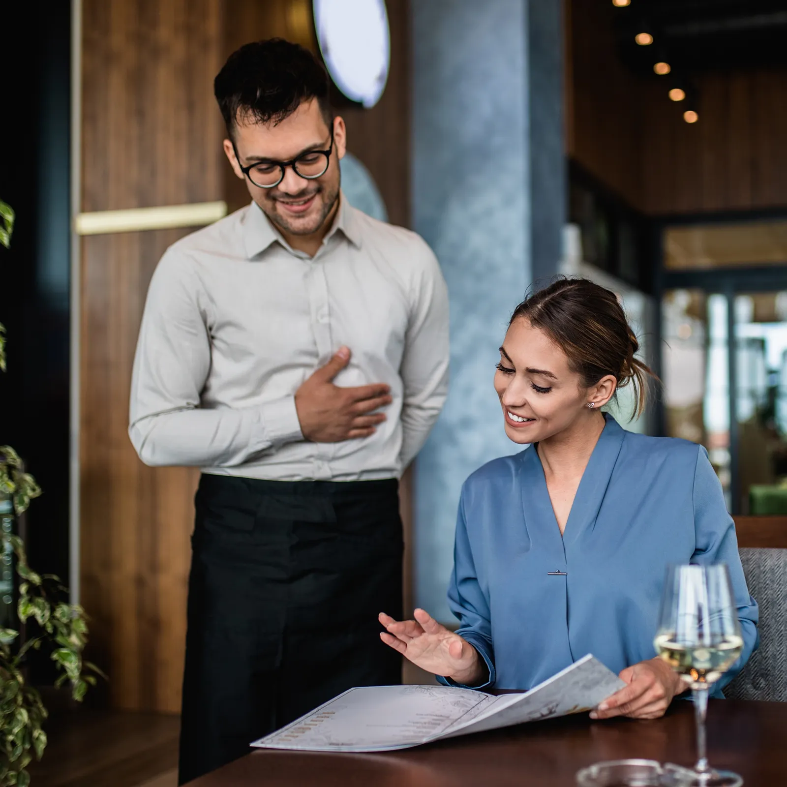 Woman smiling and pointing at menu while waiter stands attentively in restaurant.