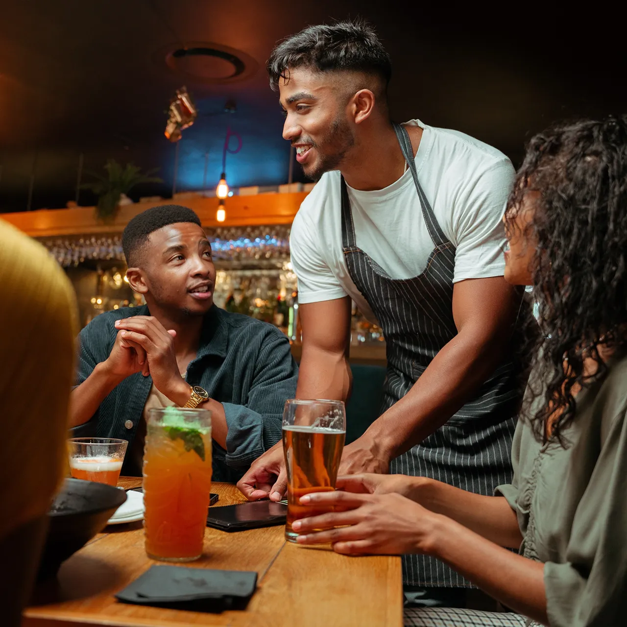 Bartender in apron serving drinks and talking to two seated customers at a bar table.
