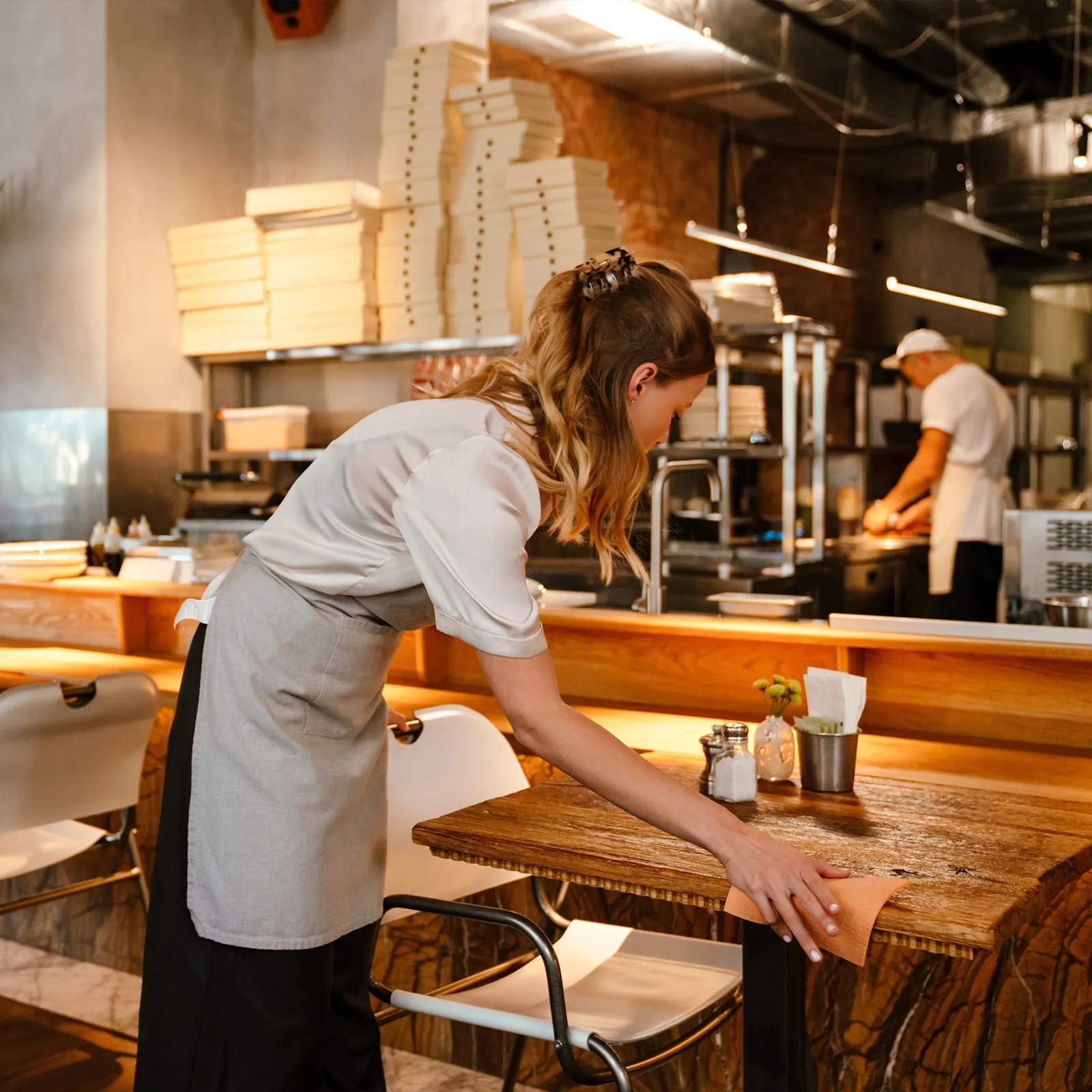 Restaurant worker wearing apron cleaning a wooden table with a cloth in a cozy restaurant.
