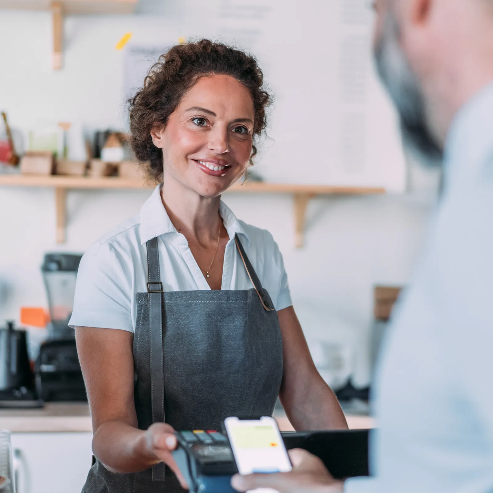Smiling female cashier in apron holding payment terminal as customer makes contactless payment.