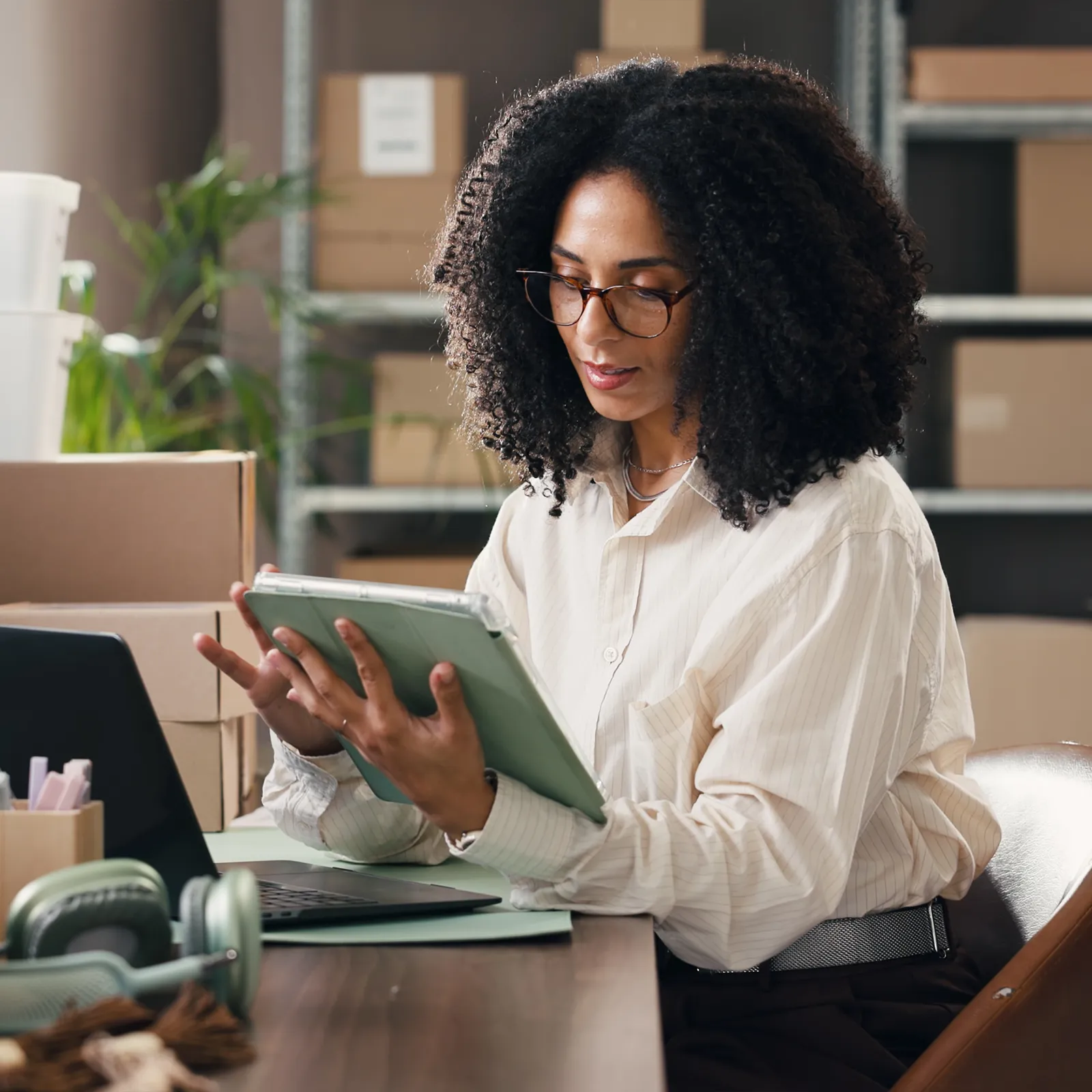 Woman with curly hair and glasses using a tablet at a desk with a laptop and boxes behind her.