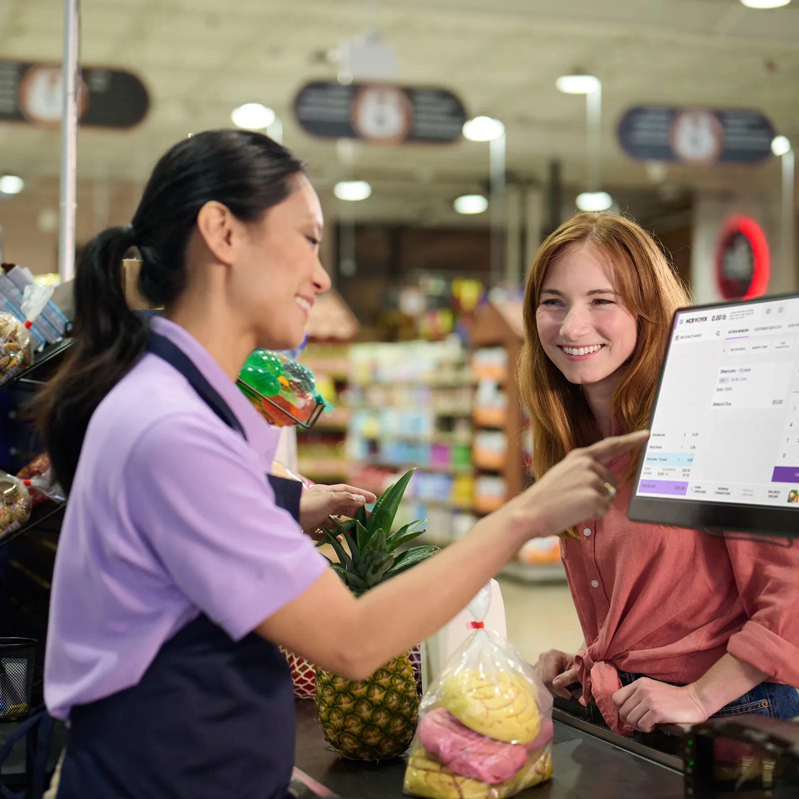 Grocery cashier in purple uniform pointing at register screen while customer smiles with groceries.