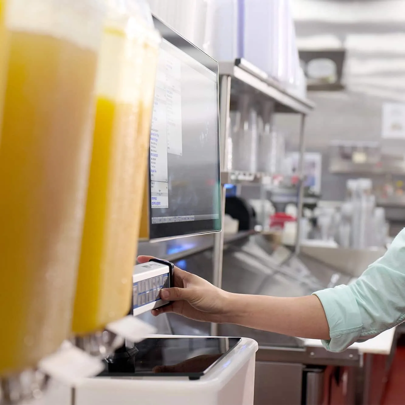 Person operating touchscreen POS system next to beverage dispensers in a commercial kitchen.