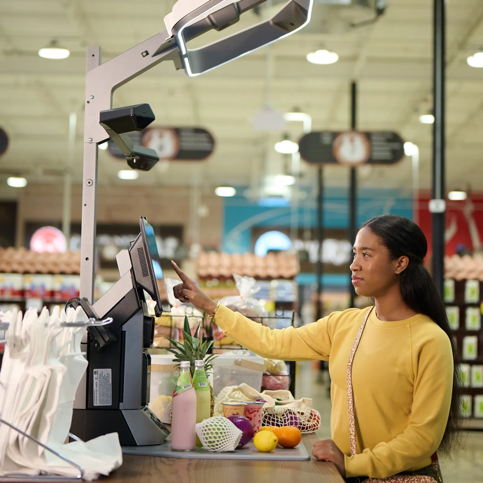 Woman in yellow sweater using self-checkout touchscreen in a grocery store with fresh produce.