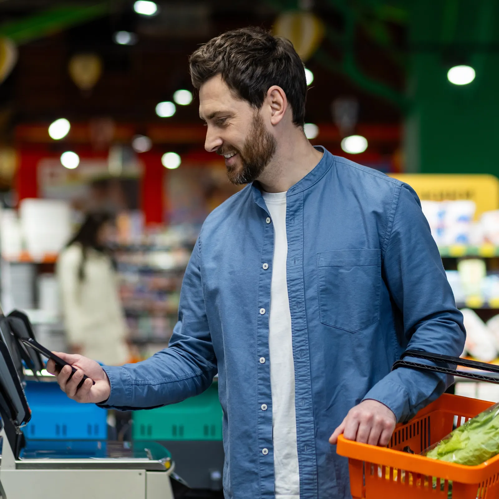 Man in blue shirt using phone to scan at a self-checkout with grocery basket in store.