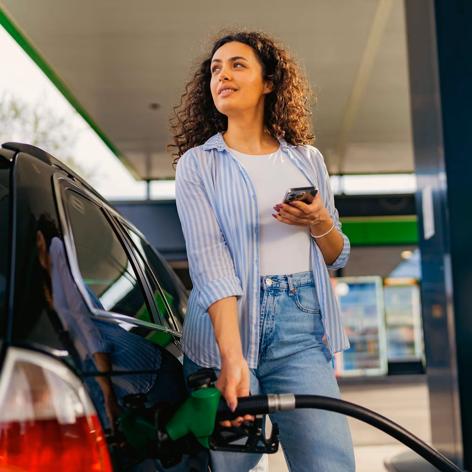 Woman holding phone while refueling her car at a gas station.