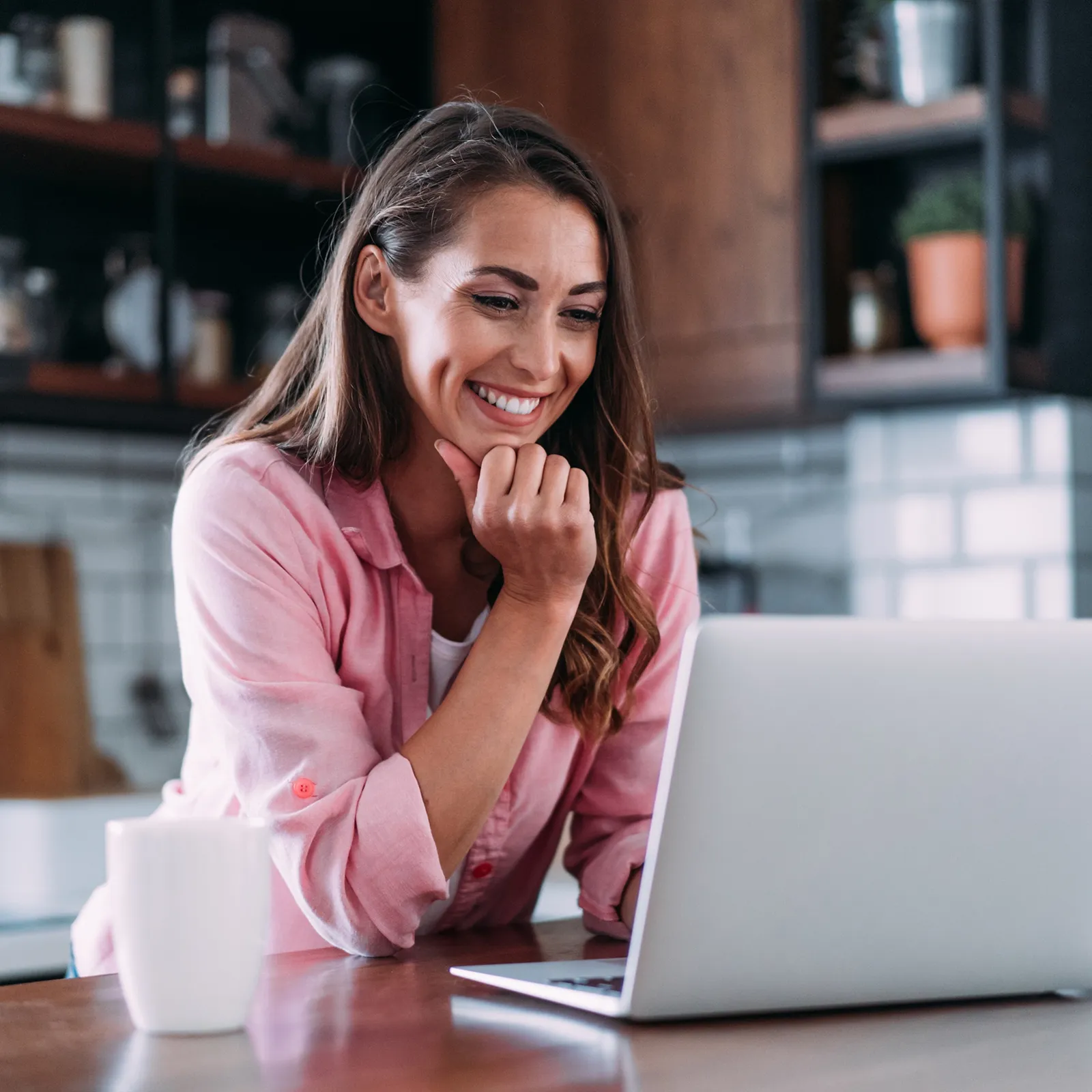 Smiling woman in pink shirt using a laptop in a modern kitchen with a coffee cup nearby.