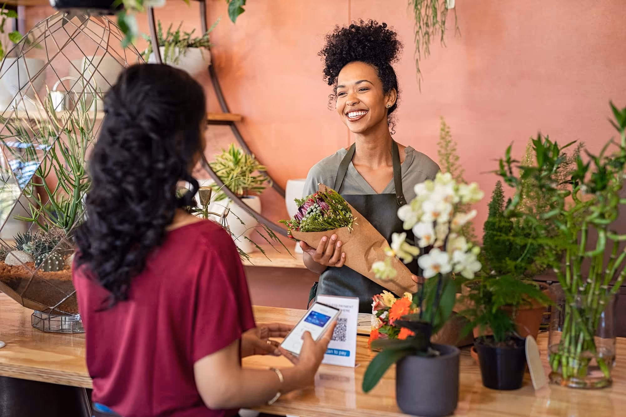 Smiling florist handing bouquet to customer using smartphone to pay at flower shop counter.