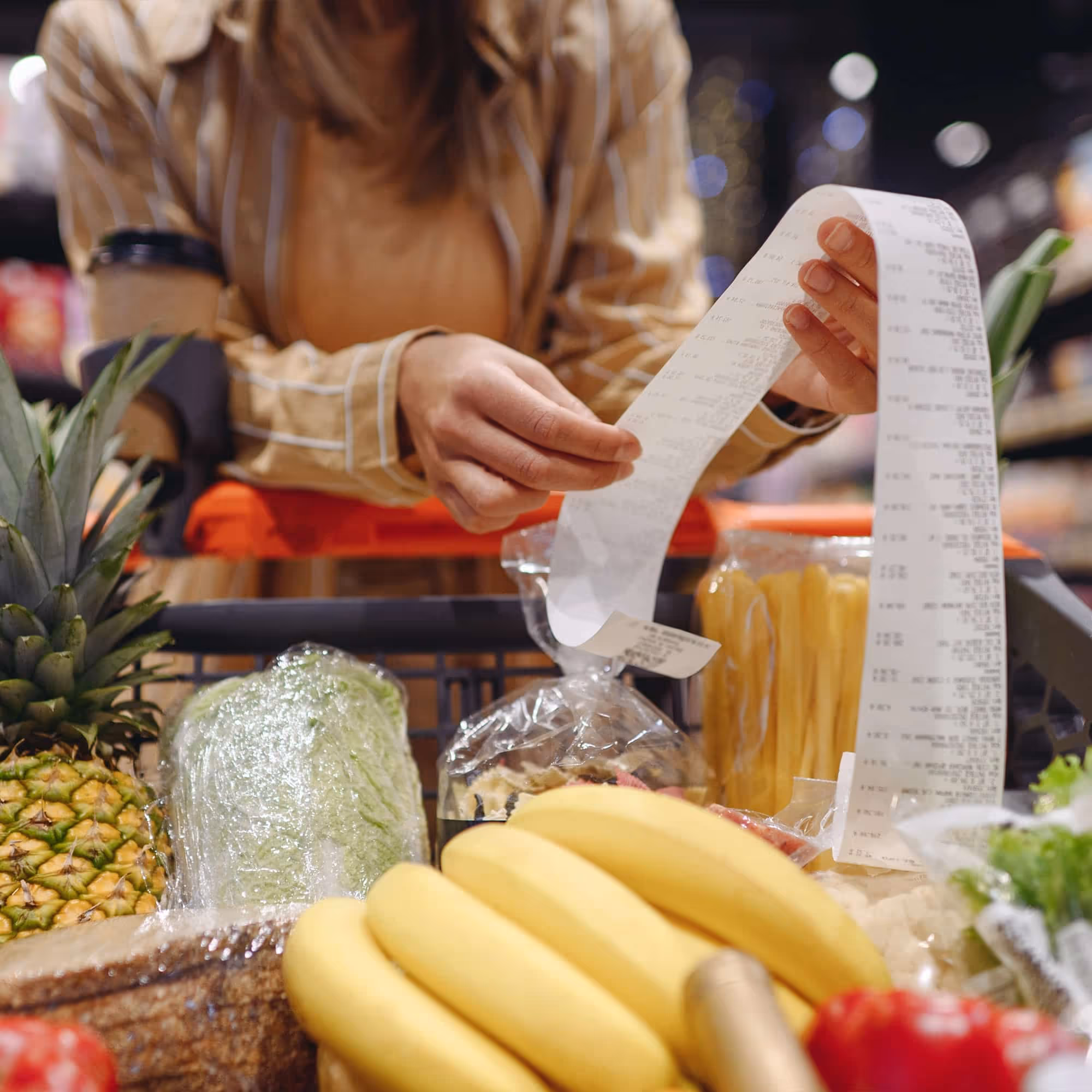 Person holding a long grocery receipt over a cart with bananas, pineapple, lettuce, and pasta.