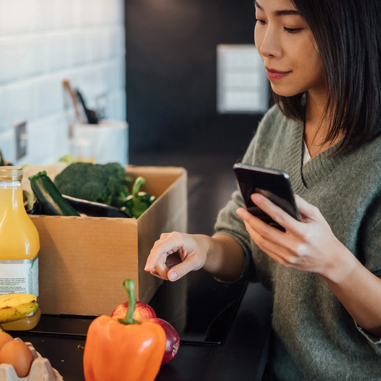 Woman holding smartphone in kitchen near fresh groceries including bell pepper, apples, and broccoli.